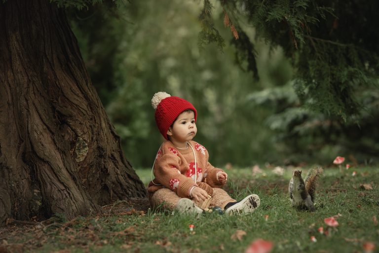 Bebé con gorro rojo sentado al pie de un árbol en el National Botanic Garden en Irlanda, junto a una ardilla en el césped.