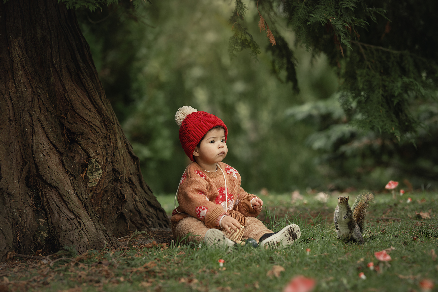 Bebé con gorro rojo sentado al pie de un árbol en el National Botanic Garden en Irlanda, junto a una ardilla en el césped.
